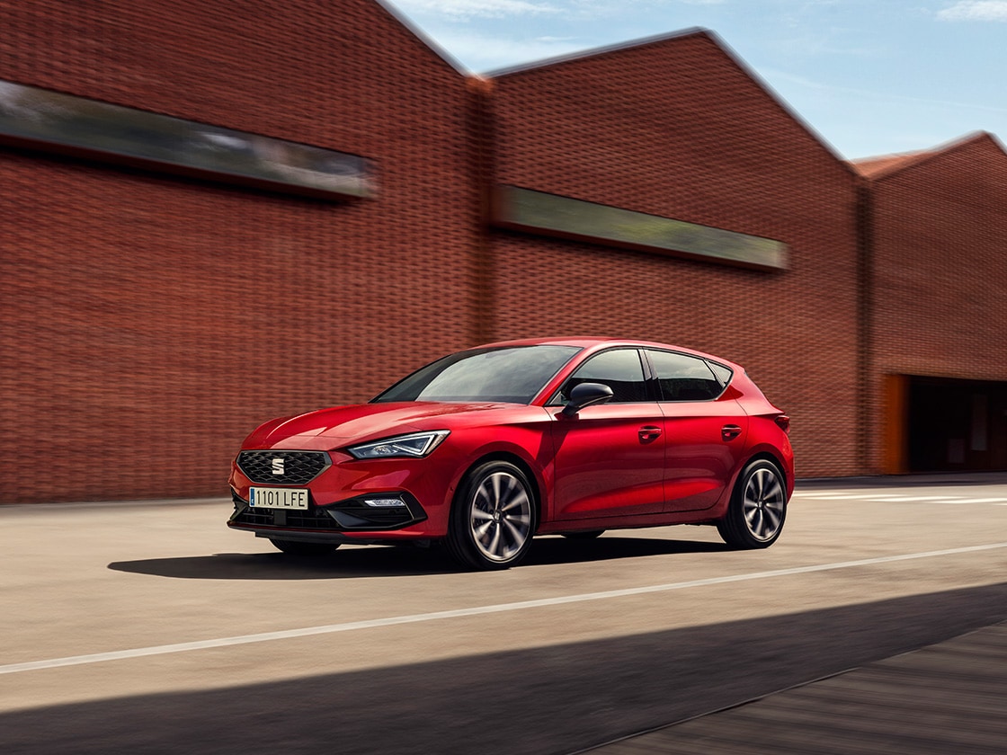 Ein roter SEAT Leon in Bewegung vor einem industriellen Hintergrund. Es gibt keine anderen Autos in der Nähe und das Fahrzeug sieht makellos aus, was die Zuverlässigkeit der Vorteile des Service- und Wartungsplans von SEAT unterstreicht.