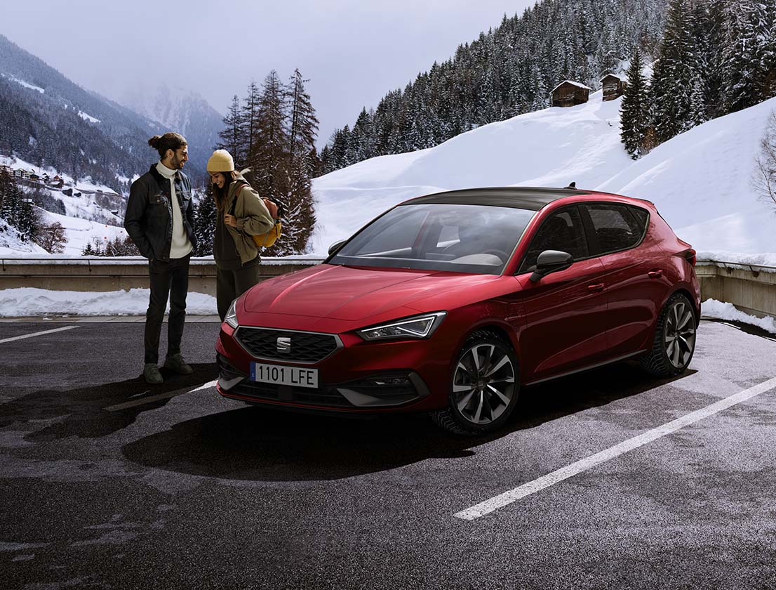 Ein Paar unterhält sich neben einem SEAT Leon in Desire Red in einer verschneiten Berglandschaft