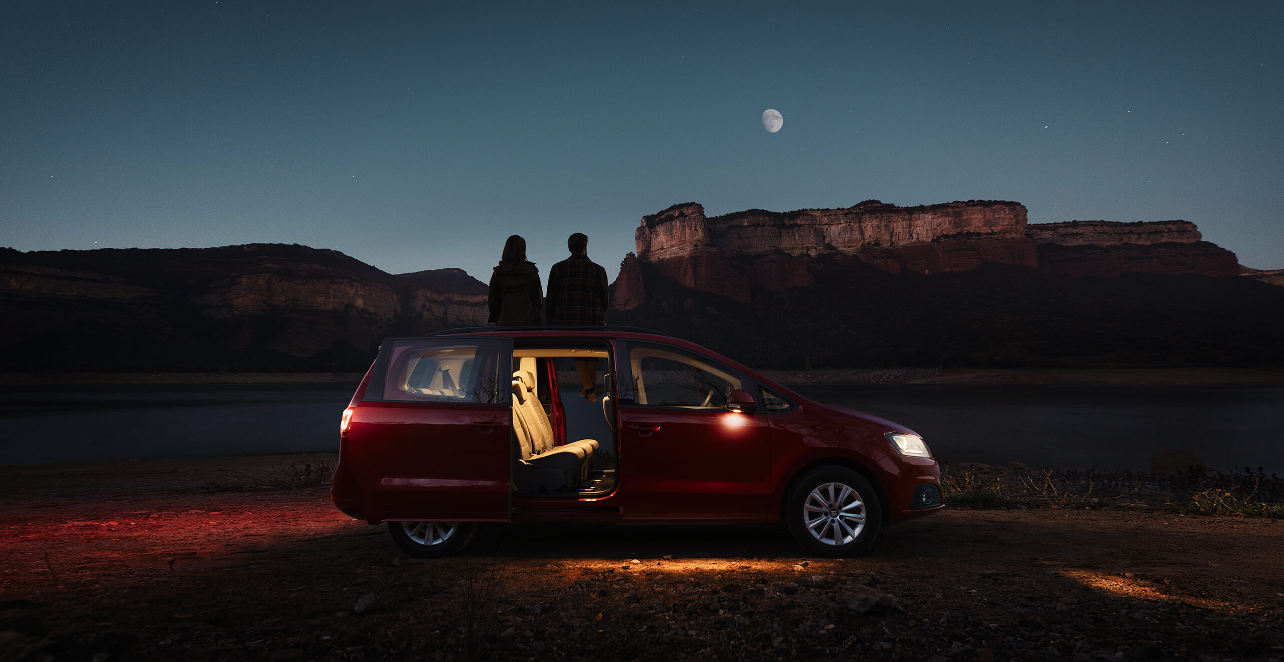 Vue latérale d'une SEAT Alhambra la nuit, les portières ouvertes, avec deux personnes regardant la lune et le paysage montagnard